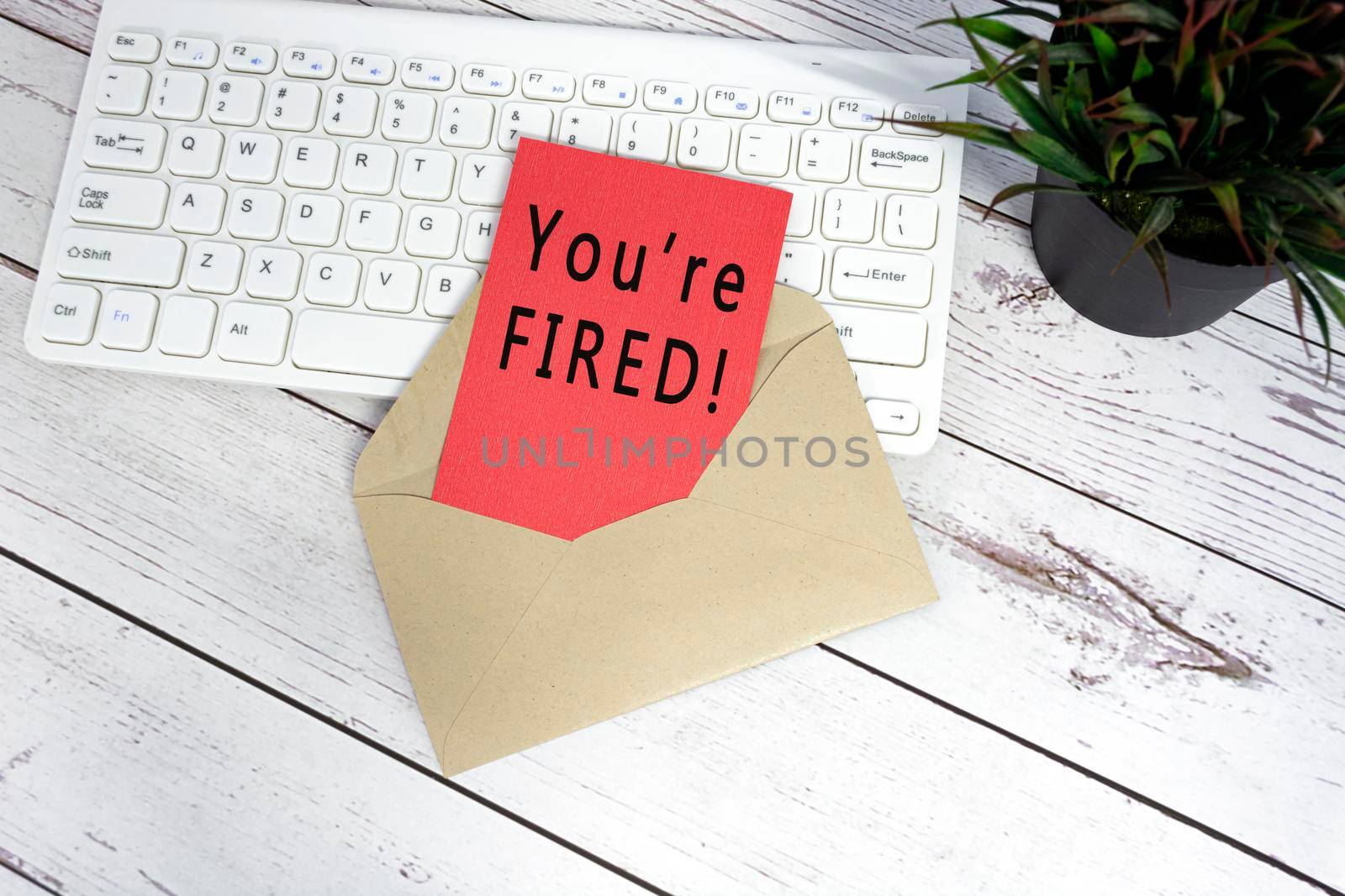 You are fired text on red note inside brown envelope with keyboard and potted plant background on wooden desk.