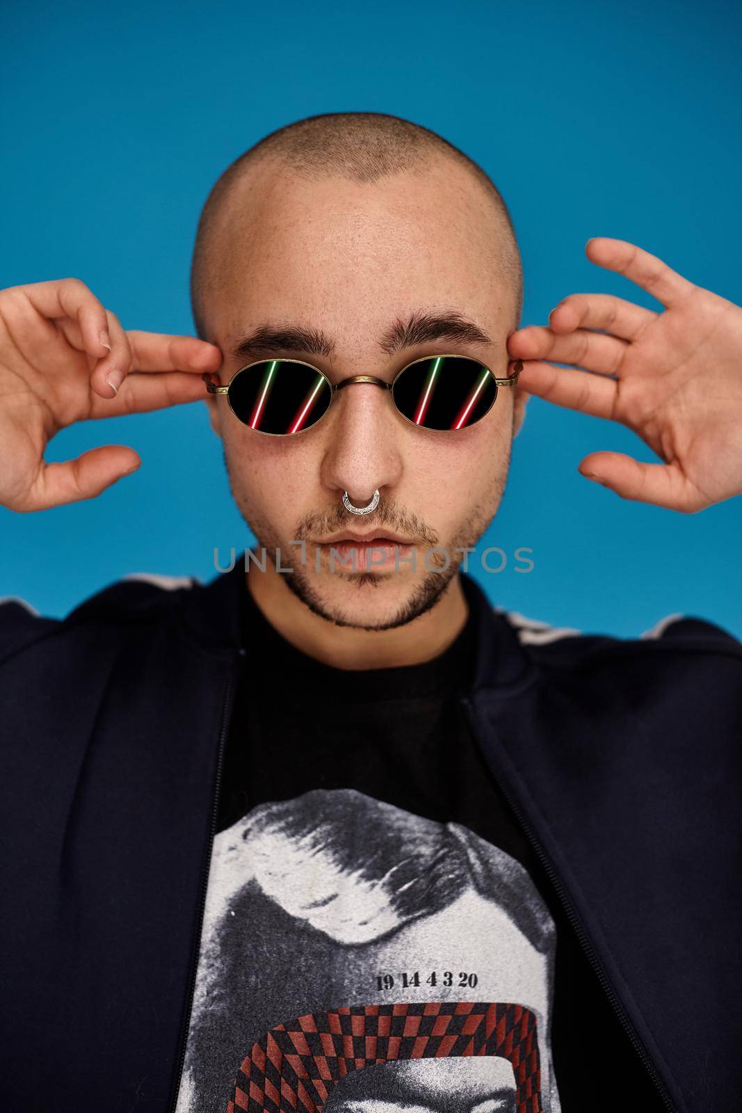 Close-up studio shot of a young tattoed bald person straightens his sunglasses, with a pirsing ring in his nose, wearing black trendy t-shirt with print and sport suit, posing against a blue background with copy space. People, style and fashion concept. 90s style