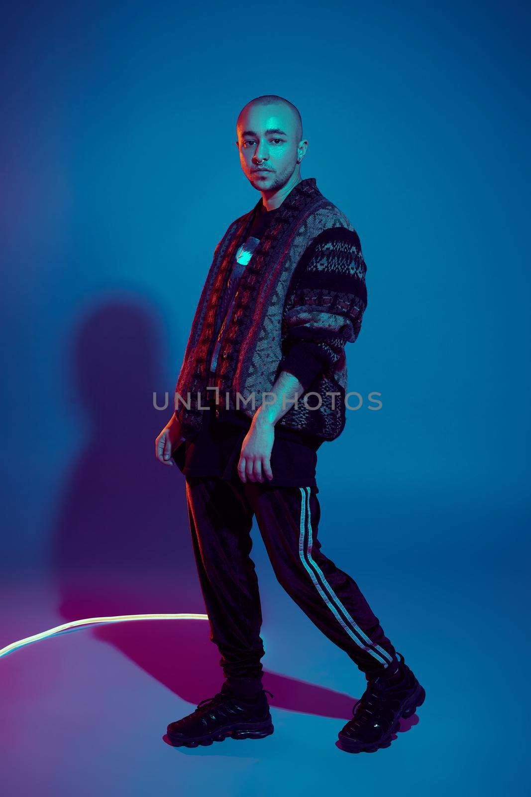 Full-length studio shot of a handsome tattoed bald guy with a pirsing ring in his nose, wearing dark jacket, black trendy t-shirt with print, sneakers and sweatpants, posing sideways against a colorful background with copy space. People, style and fashion concept. 90s style