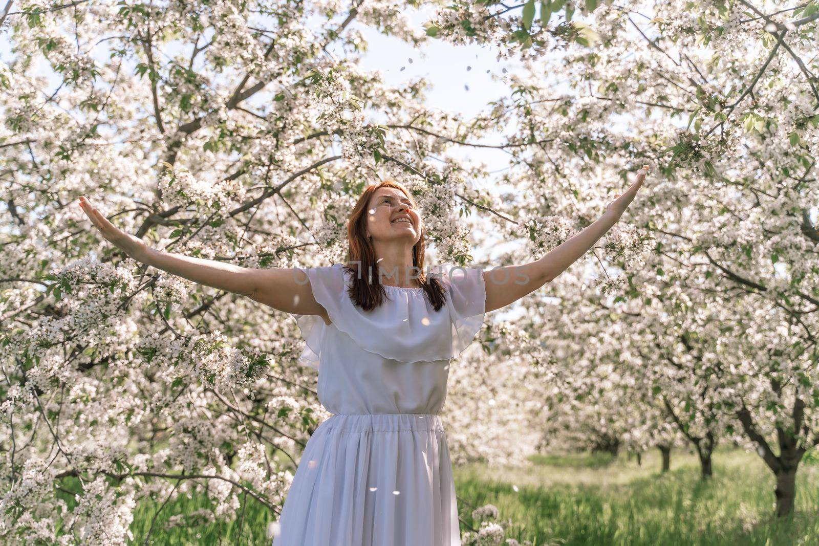 Fantasy woman in long white elegant fashion long dress walks in green spring blossom cherry garden. Happy cheerful girl princess bride. Skirt fabric flies flowing waving in wind motion