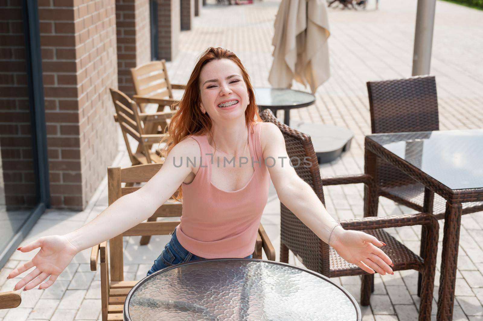 Young red-haired woman sits in a street cafe and stretches her arms to hug