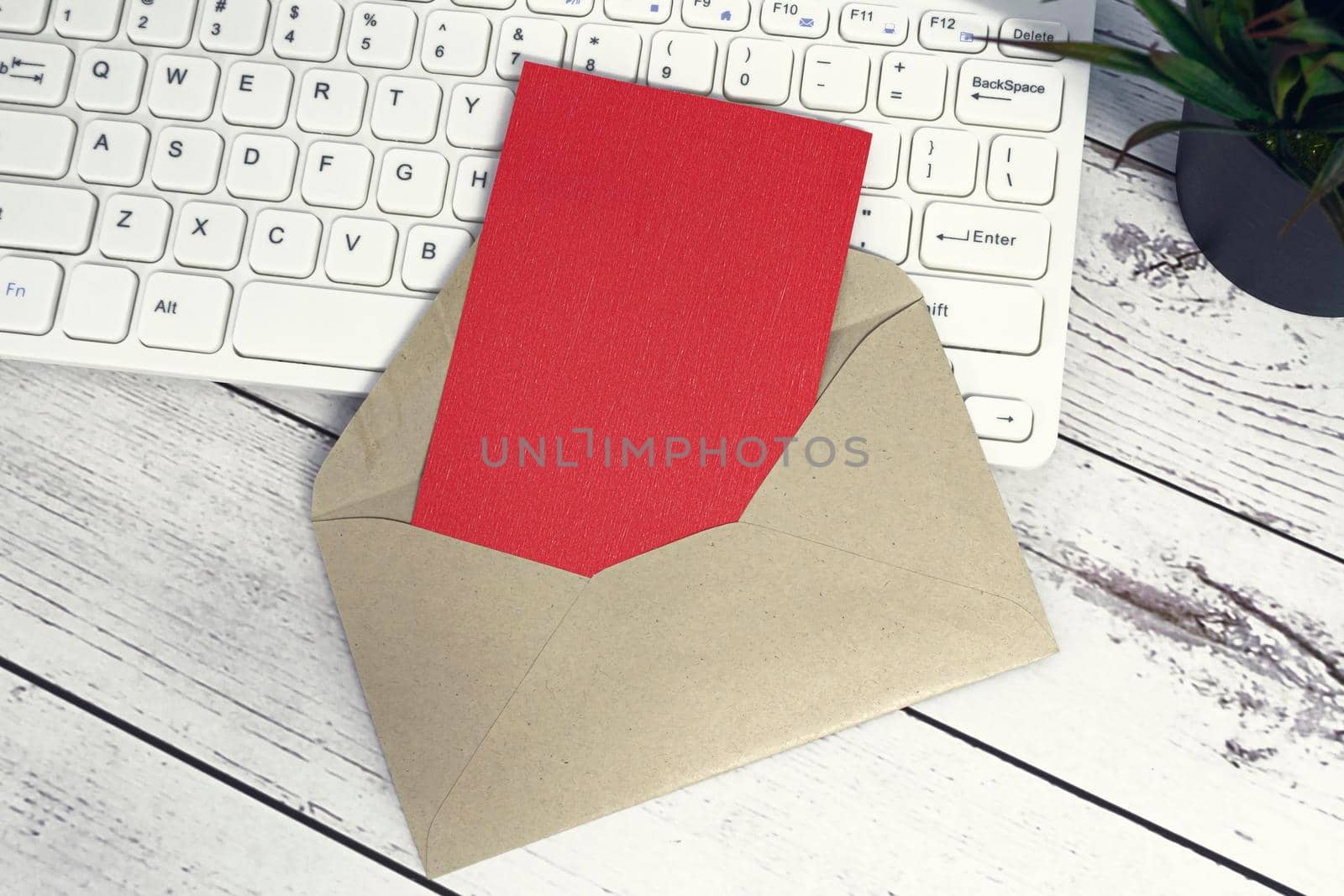 Red note inside brown envelope with keyboard and potted plant background on wooden desk. Copy space. Flat lay.