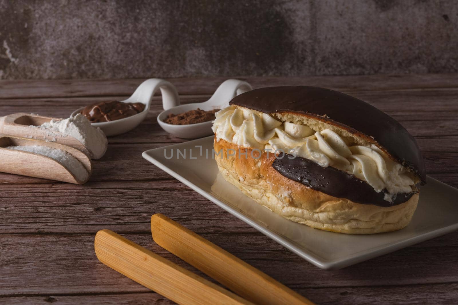 chocolate-covered cream-filled bun on a white plate with the ingredients and wooden tongs.
