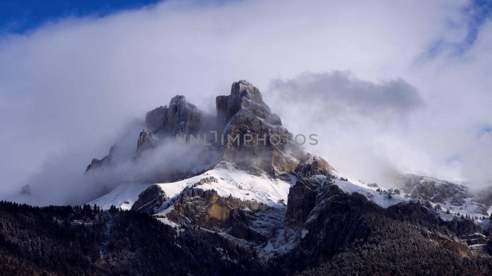 Photograph of sunlight and clouds obscuring and revealing the jagged peak of a mountain in the French Alps mountain range.