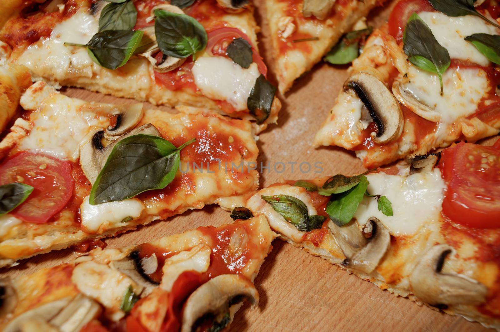 Close-up of an appetizing, freshly baked vegetarian pizza with addition ripe juicy tomatoes, organic champignons and aromatic basil herbs, divided into portions on a wooden background. Selective focus