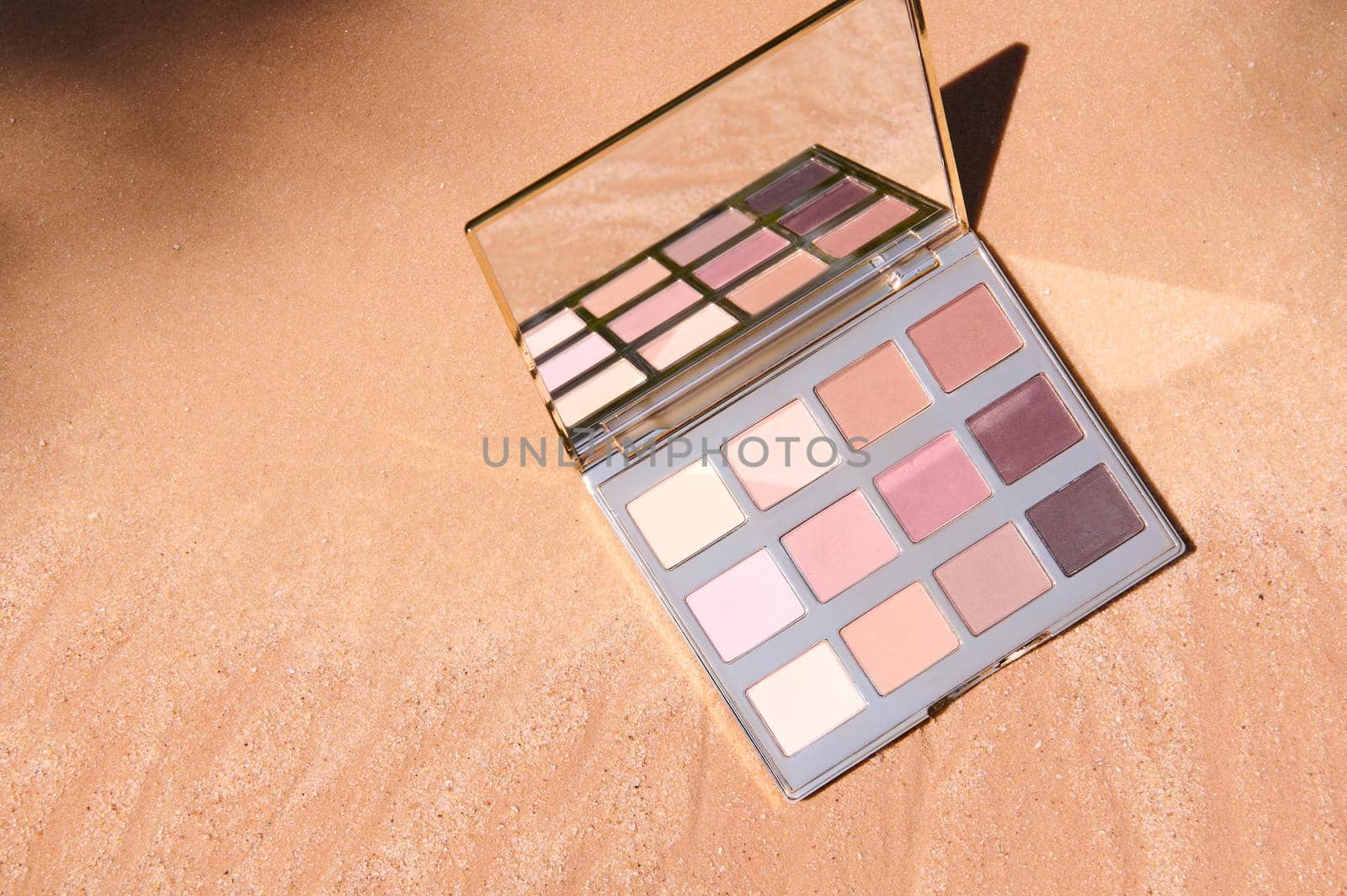 Top view of a beautiful luxurious makeup palette of eyeshadows with gradient from dark brown to shades of beige color, reflecting in cosmetic mirror, on golden sandy background with copy ad space.