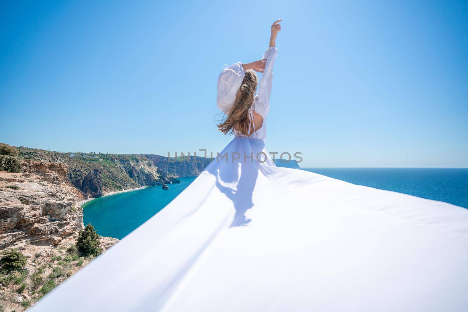 Blonde with long hair on a sunny seashore in a white flowing dress, rear view, silk fabric waving in the wind. Against the backdrop of the blue sky and mountains on the seashore