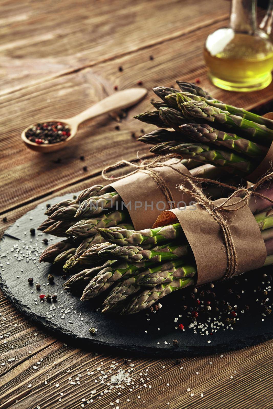 Bunches of an edible, organic stalks of asparagus on a stone slate, wooden background. Fresh, green vegetables with olive oil and seasonings, wooden spoon, top view. Healthy meal. Fall harvest, agricultural farming concept.