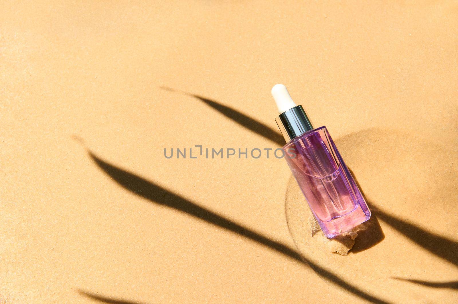 Liquid cosmetic product with collagen and peptides in light purple glass bottle with dropper on golden sandy background and shadow from palm leaf. Skincare essence for beautiful healthy skin. Top view