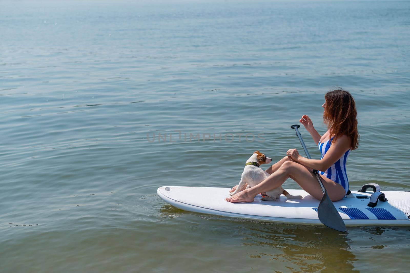 A woman is riding a sup surfboard with a dog on the lake. The girl goes in for water sports with her pet