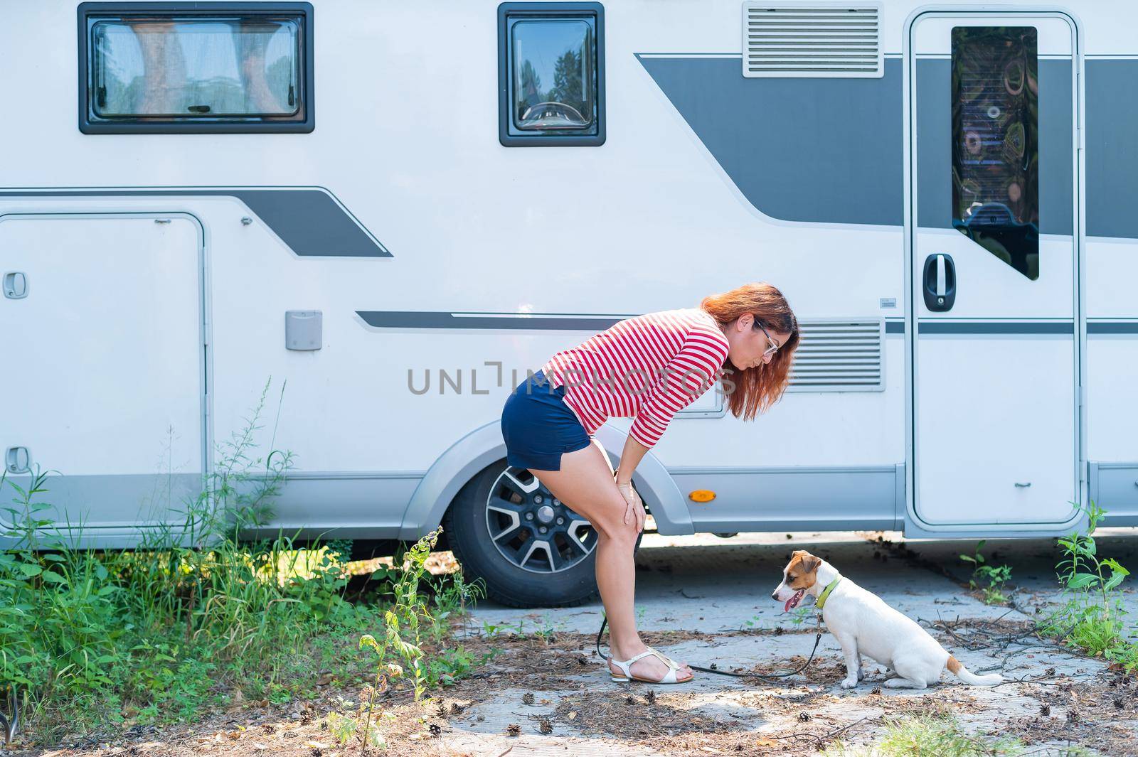 Beautiful caucasian woman is training a dog next to the motorhome. Travel with your pet in a mobile RV. Red-haired girl playing with Jack Russell Terrier. Female trailer driver. by mrwed54