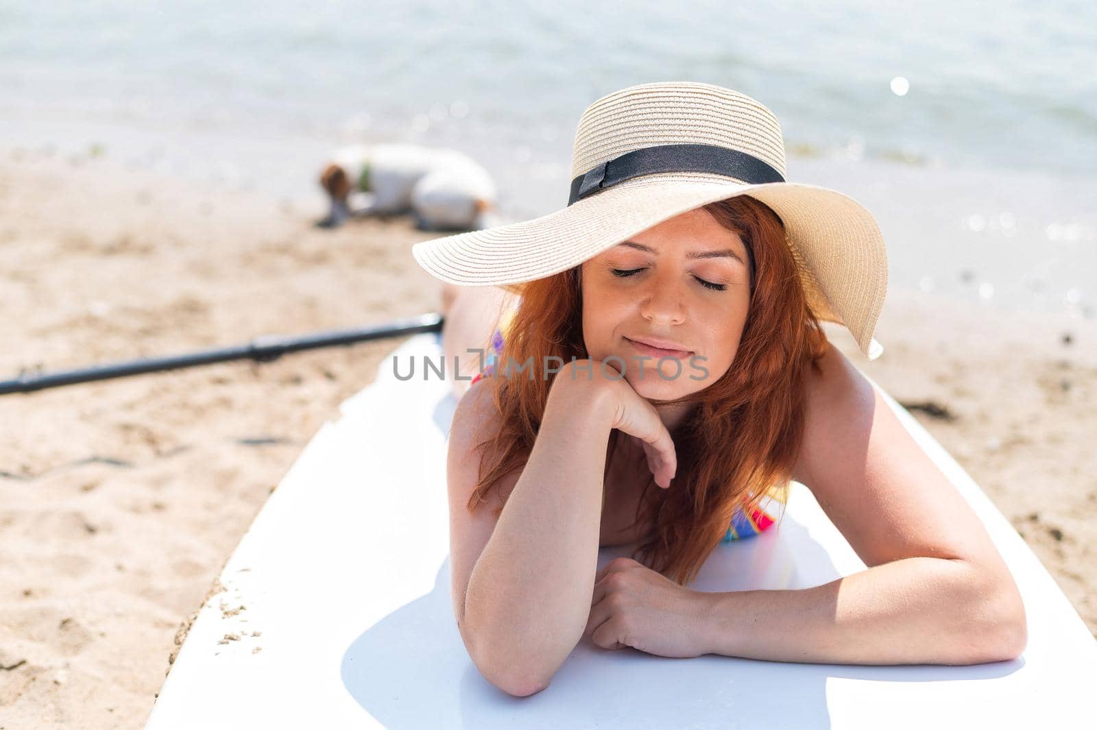 Smiling red-haired woman wearing a hat and swimsuit lies on a paddle board. Water sports. Active holidays. by mrwed54