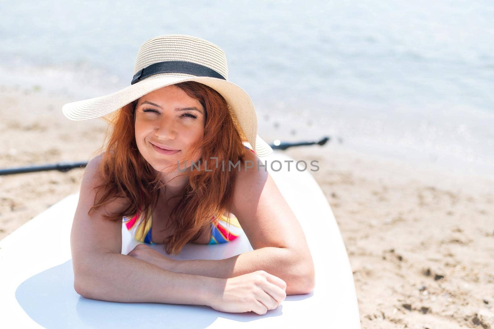 Smiling red-haired woman wearing a hat and swimsuit lies on a paddle board. Water sports. Active holidays