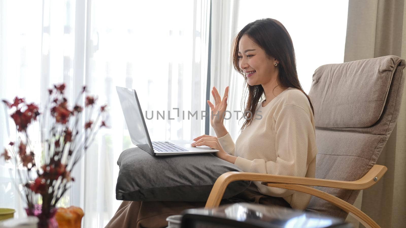 Positive young woman waving hand, chatting online, making video call on her laptop by prathanchorruangsak