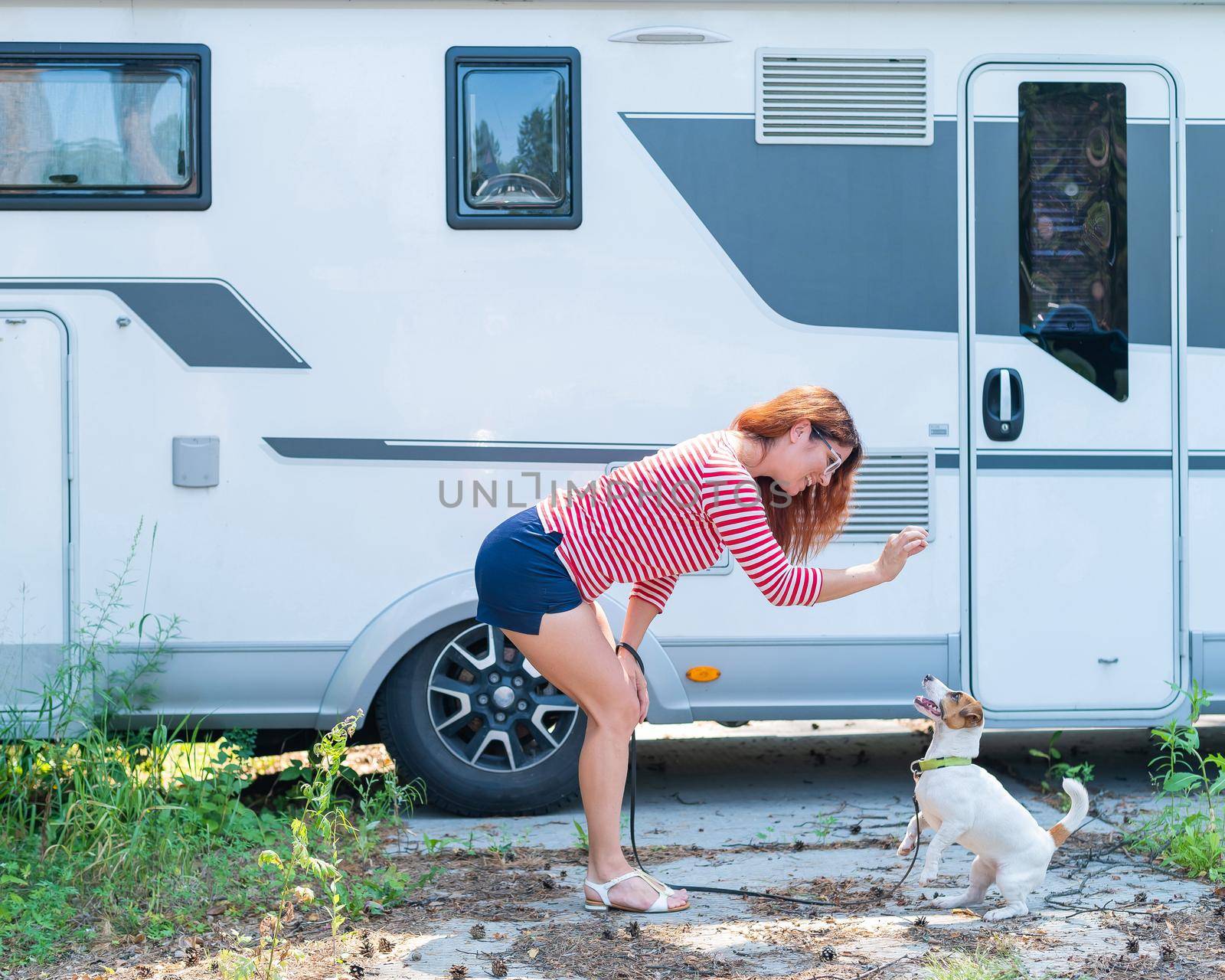 Beautiful caucasian woman is training a dog next to the motorhome. Travel with your pet in a mobile RV. Red-haired girl playing with Jack Russell Terrier. Female trailer driver. by mrwed54