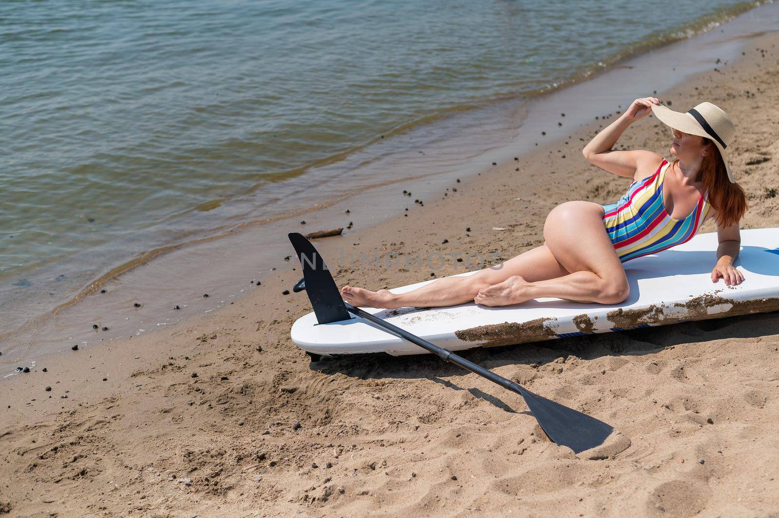 A woman in a swimsuit and a hat is posing lying on a sup board on the beach