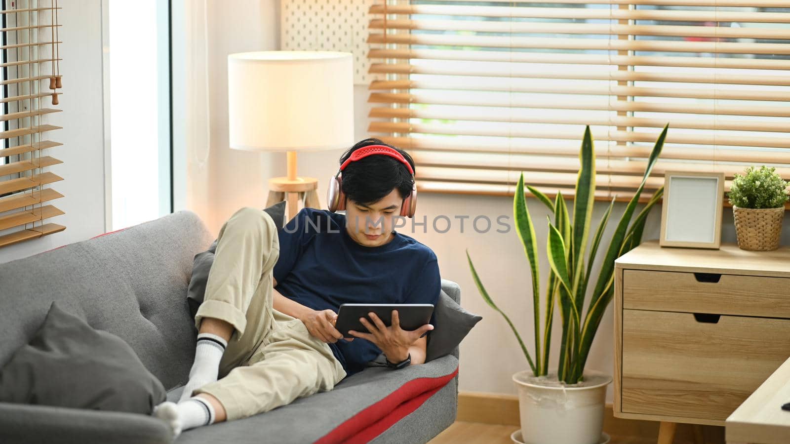 Relaxed asian man wearing wireless headphone lying on couch and surfing internet on digital tablet.
