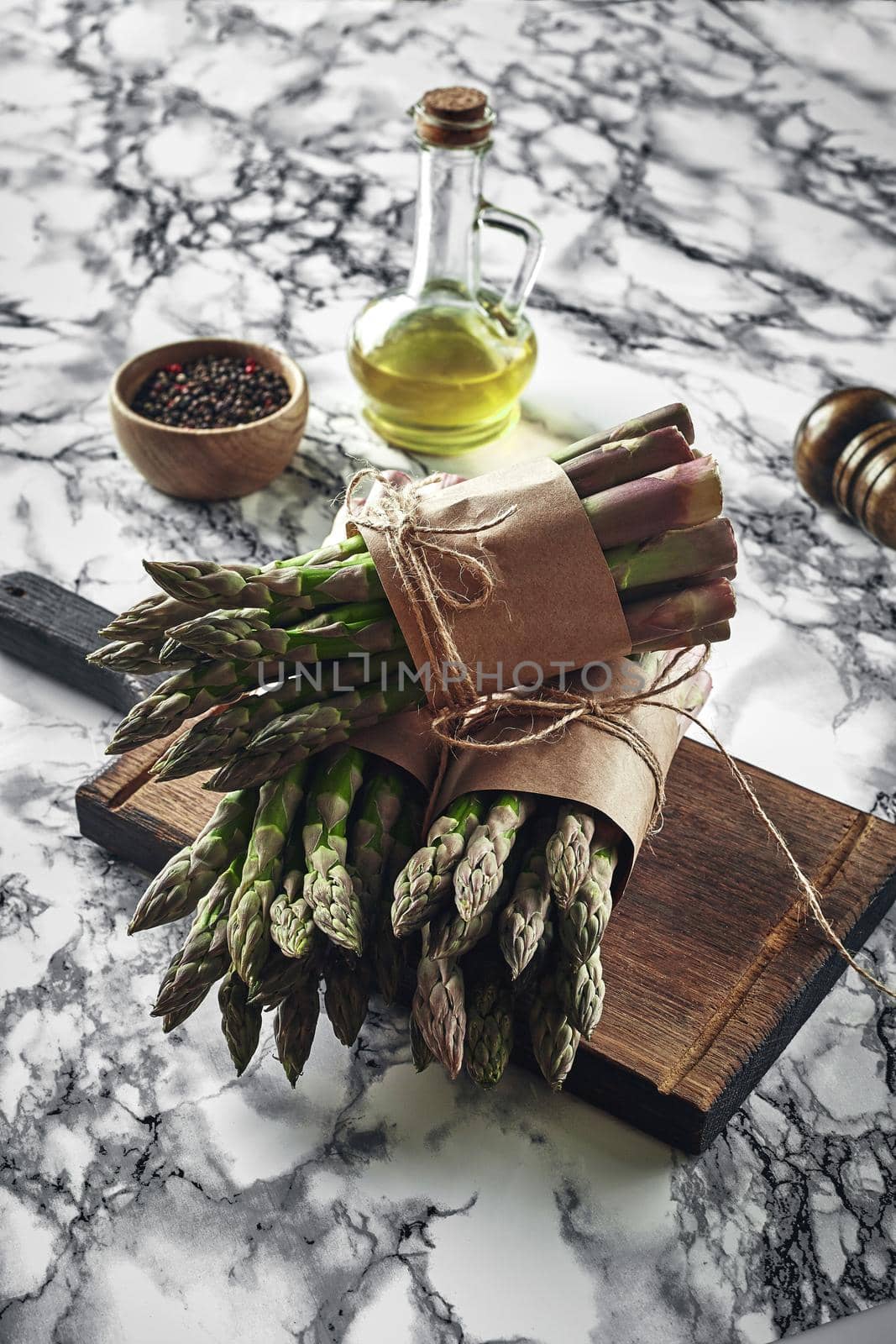 Bunch of an edible, fresh stems of asparagus on a wooden board, marble background. Green vegetables with olive oil and seasonings, top view. Healthy eating. Spring harvest, agricultural farming concept.