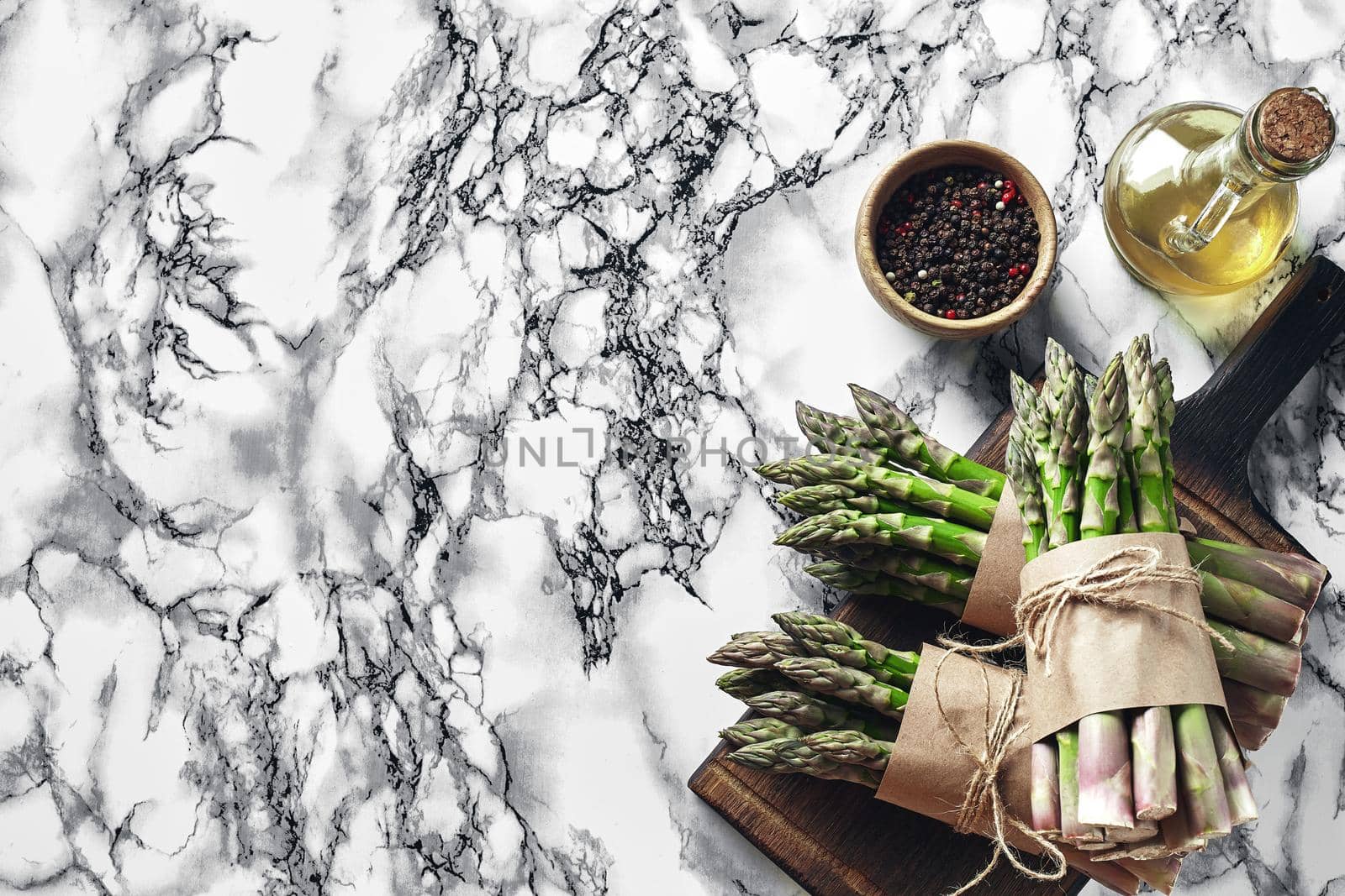 Bunch of an edible, fresh sprouts of asparagus on a wooden board, marble background. Green vegetables with olive oil and seasonings, top view. Healthy food. Spring harvest, agricultural farming concept.