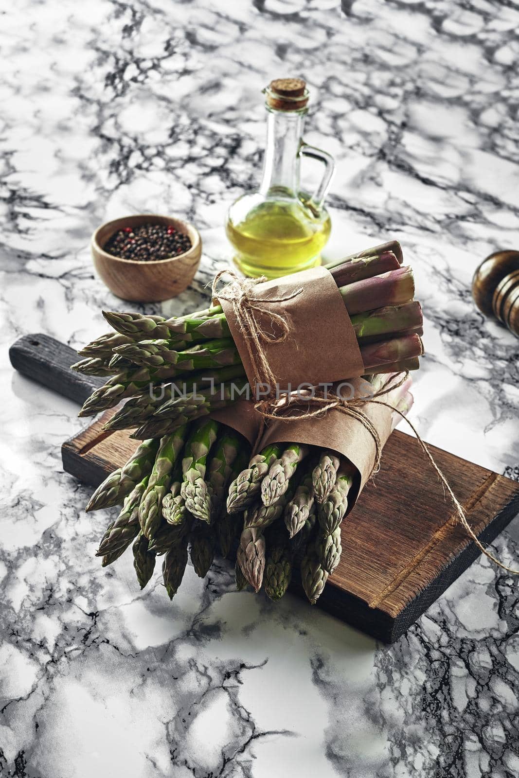 Bunch of an edible, green sprigs of asparagus on a wooden board, marble background. Fresh vegetables with olive oil and seasonings, top view. Healthy eating. Spring harvest, agricultural farming concept.