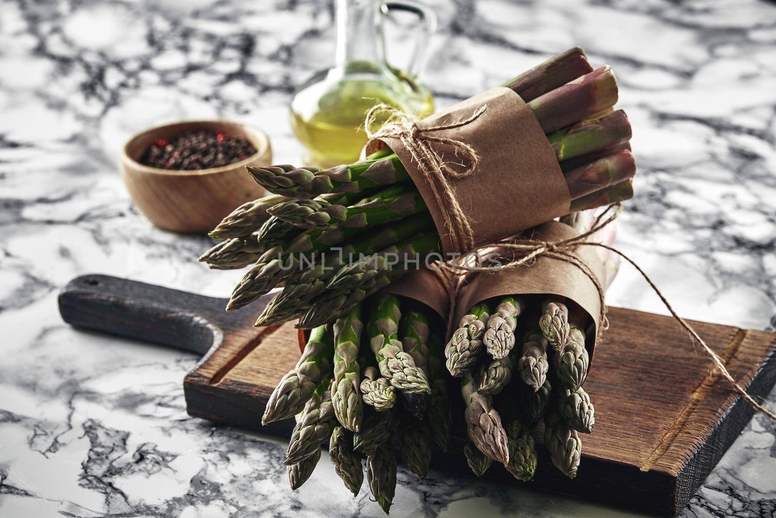 Bunch of an edible, green spears of asparagus on a wooden board, marble background. Fresh vegetables with olive oil and seasonings, top view. Healthy eating. Spring harvest, agricultural farming concept.