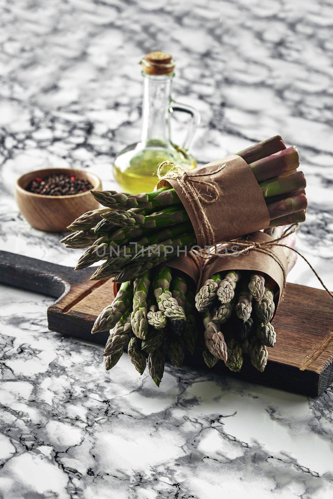 Bunch of an edible, green stalks of asparagus on a wooden board, marble background. Fresh vegetables with olive oil and seasonings, top view. Healthy meal. Spring harvest, agricultural farming concept.