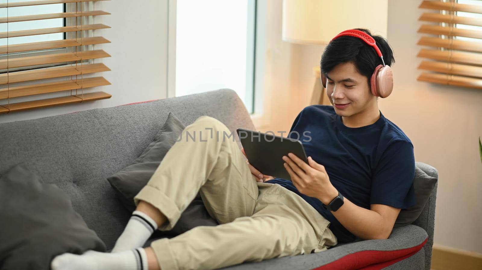 Satisfied young asian man using digital tablet, chatting in social networks while sitting on couch in cozy living room.