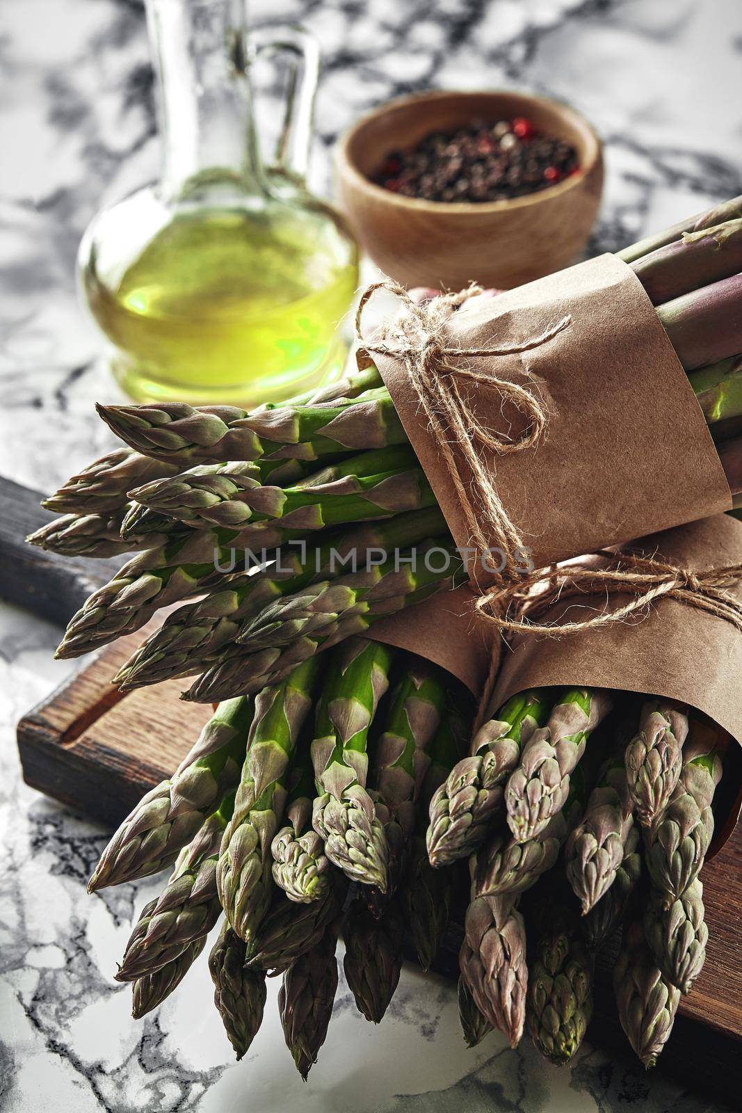 Bunch of an edible, ripe stalks of asparagus on a wooden board, marble background. Fresh, green vegetables with olive oil and seasonings, top view. Healthy eating. Spring harvest, agricultural farming concept.