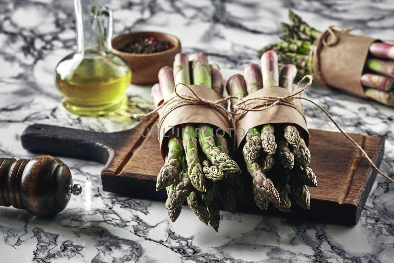 Bunch of an edible, ripe stems of asparagus on a wooden board, marble background. Fresh, green vegetables with olive oil and seasonings, top view. Healthy eating. Spring harvest, agricultural farming concept.
