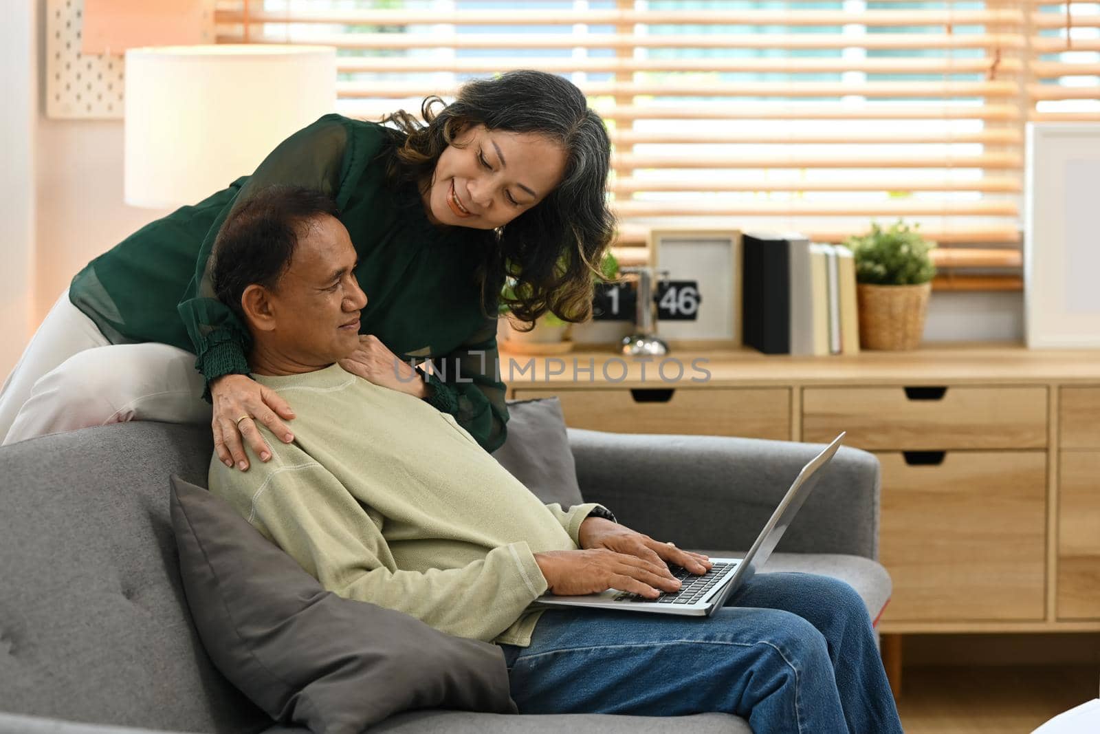 Loving senior couple resting on couch in living room and using laptop. Elderly and technology concept.