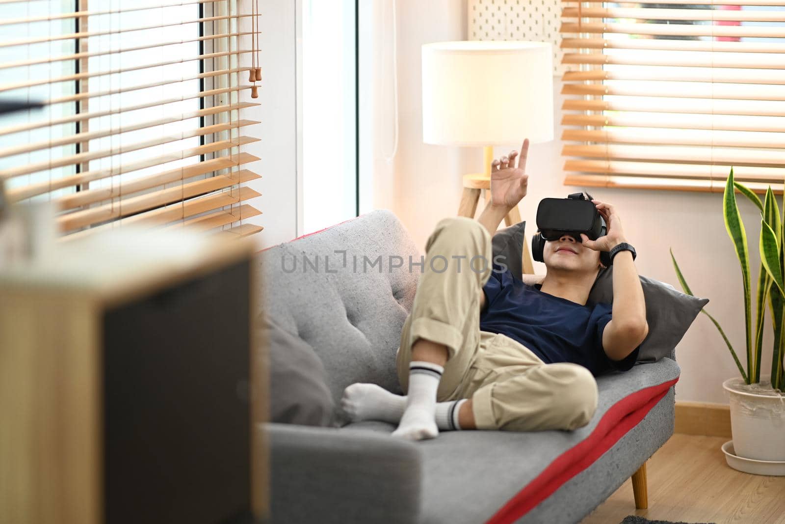Young man in casual clothes lying on couch and watching movie or play video game on virtual reality headset by prathanchorruangsak