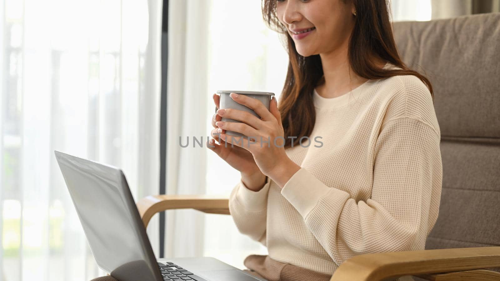 Smiling asian woman in white sweater holding coffee cup and using laptop in cozy living room.
