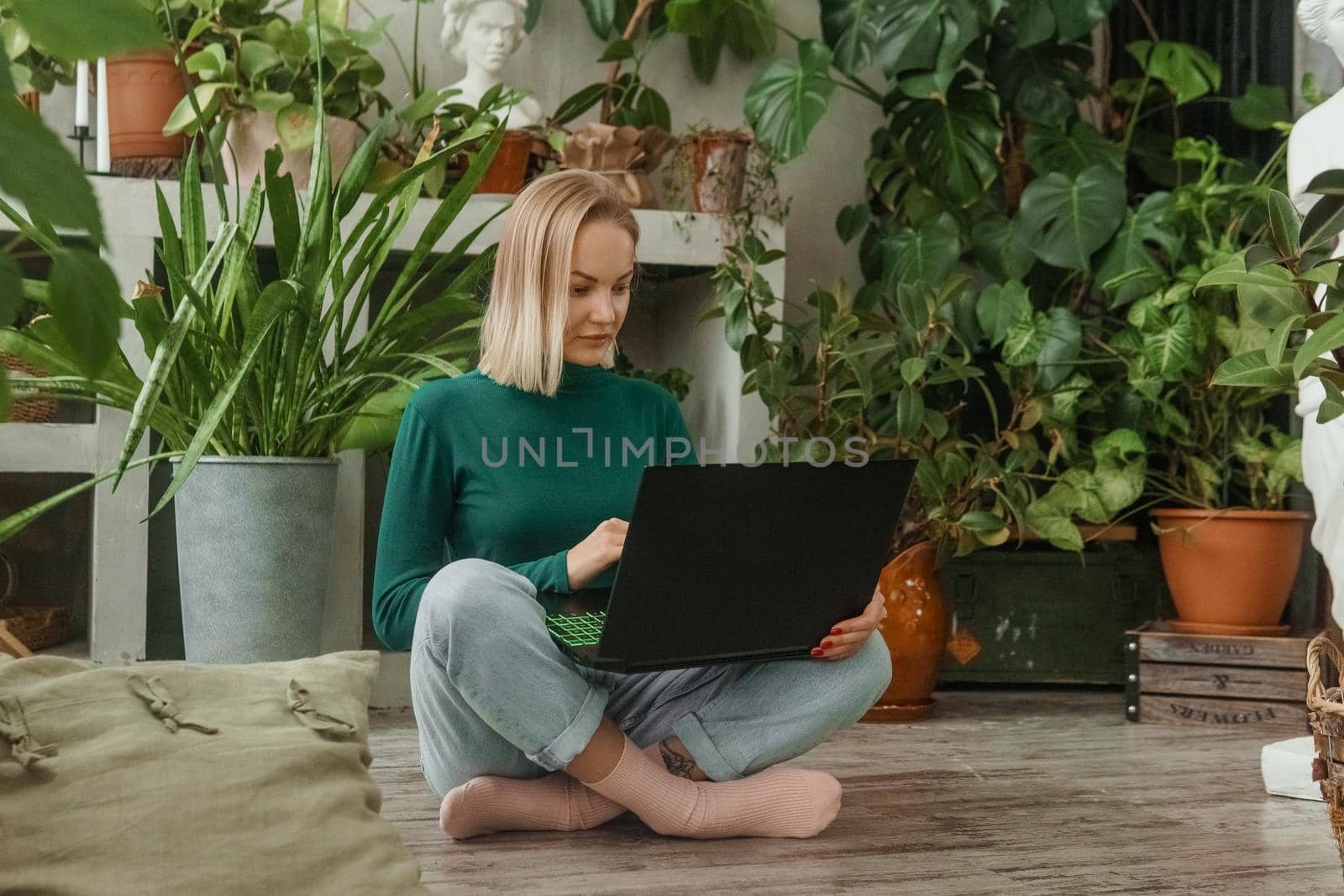 A blonde woman in a room with a lot of green indoor plants is working on a laptop. The concept of biophysical design in the interior. Work from home, work as a freelancer by Annu1tochka