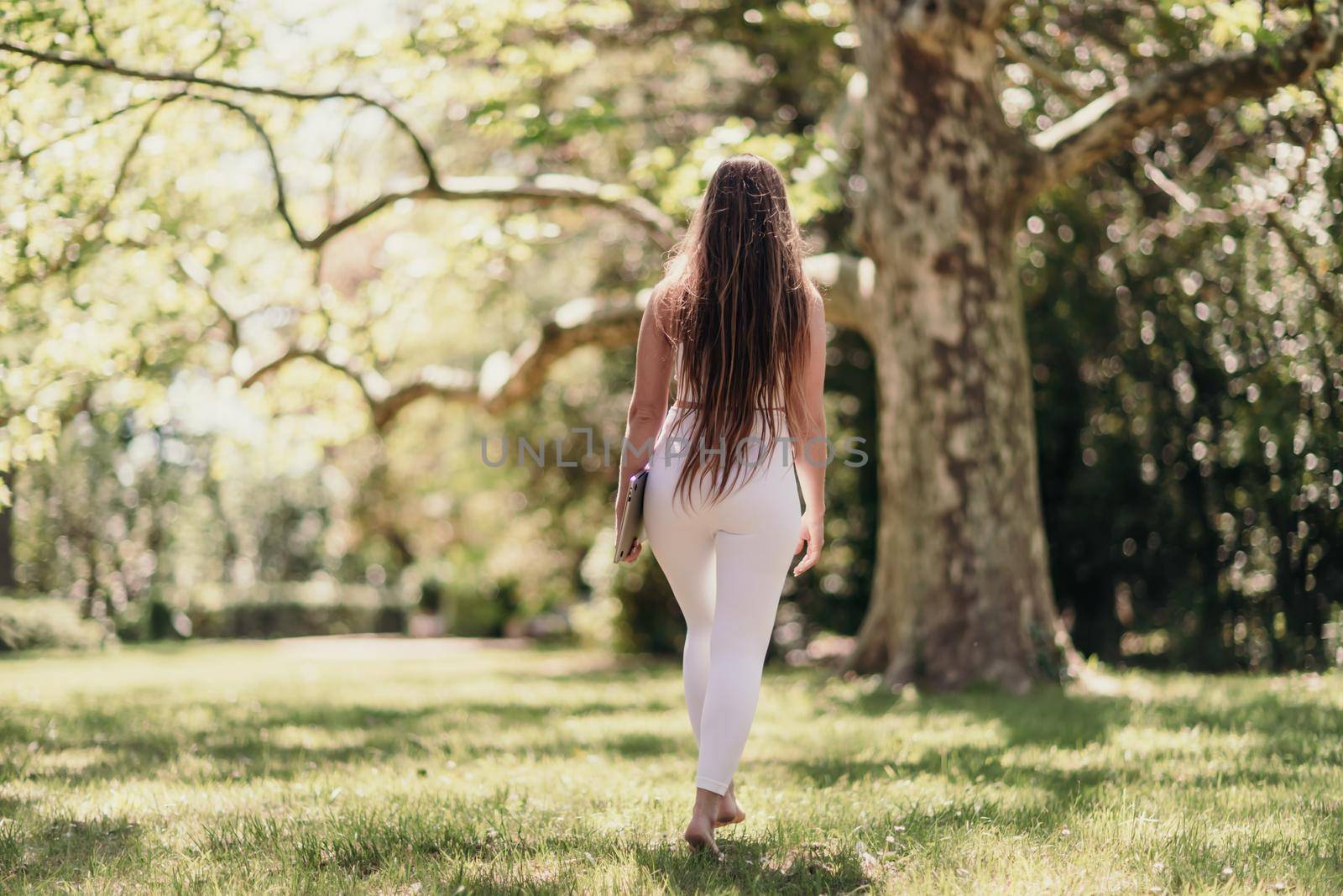 back view of a beautiful woman in a white tracksuit walking in the park with a laptop.