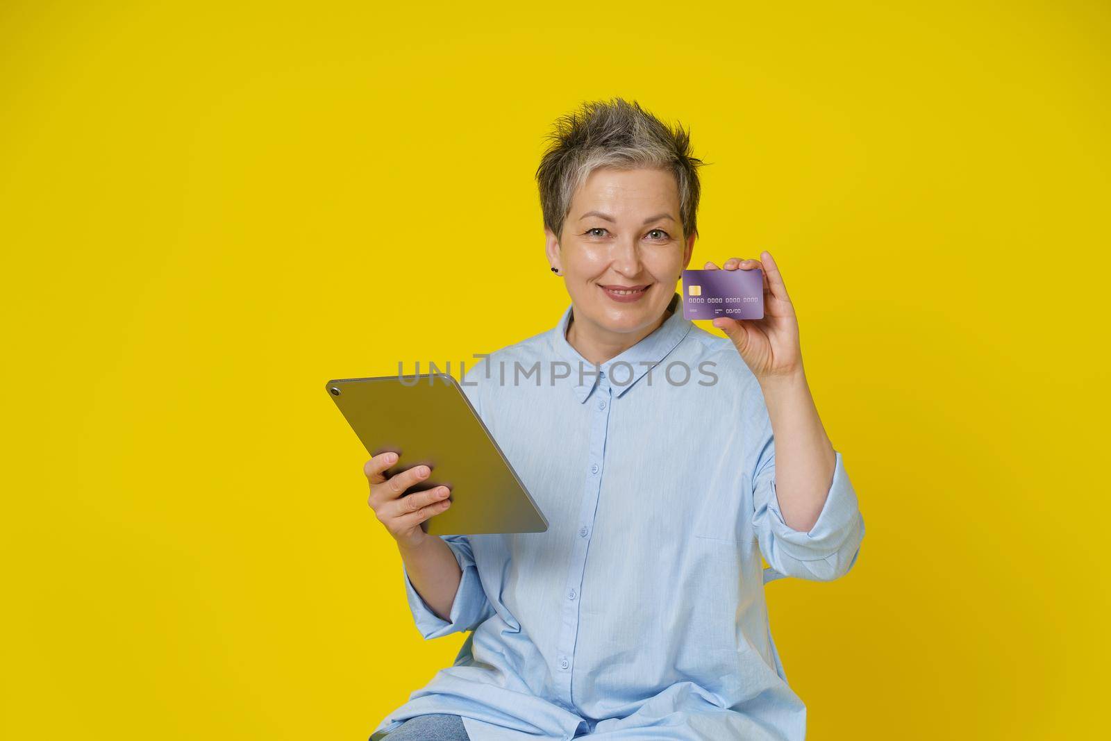 Mature woman with grey hair holding credit, debit card and tablet pc in hand making online payment or shopping online, isolated on yellow background. E-commerce, online banking concept.