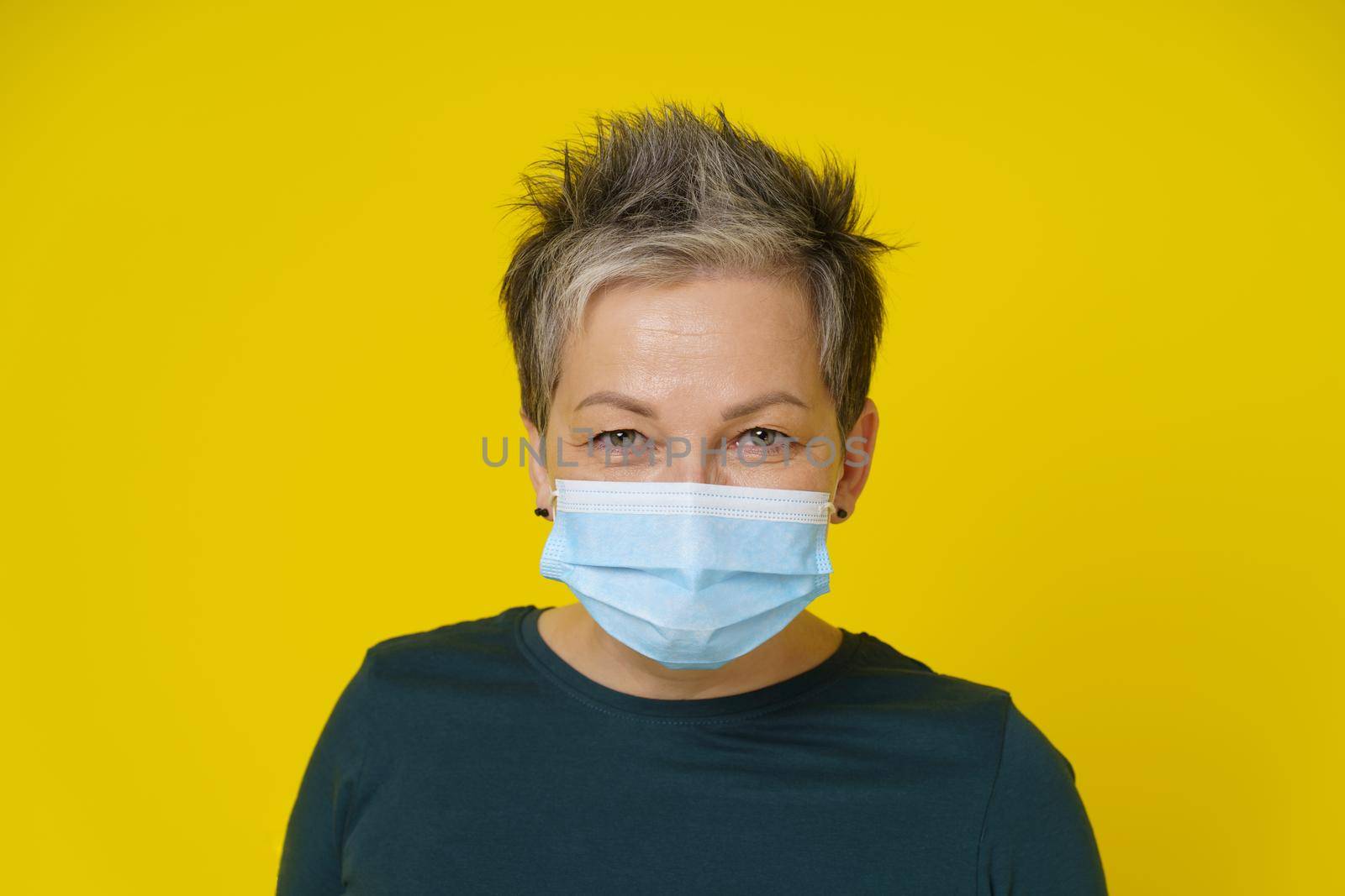 Mature woman wearing medical face mask with happy eyes smiling looking at camera wearing green blouse isolated on yellow background. Close up adult woman in medical mask.