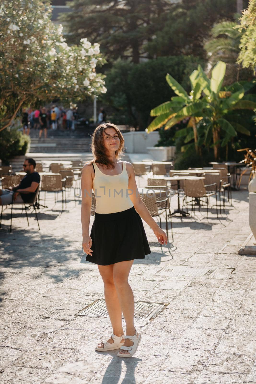 Girl Tourist Walking Through Ancient Narrow Street On A Beautiful Summer Day In MEDITERRANEAN MEDIEVAL CITY, OLD TOWN BUDVA, MONTENEGRO. Young Beautiful Cheerful Woman Walking On Old Street At Tropical Town. Pretty Girl Looking At You And Smiling by Andrii_Ko