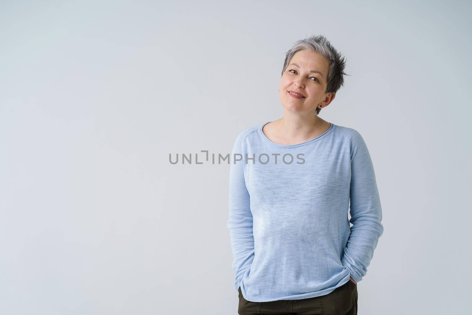 Happy dreamy mature grey haired woman 50s posing tenderly looking at camera with hands in pockets, copy space on left isolated on white background. Mature people healthcare.