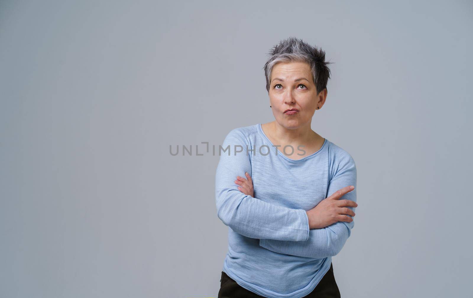 Grey haired mature woman standing with arms folded and wondering face expression in blue blouse copy space on left isolated on white background. Healthcare concept. Aged beauty concept. Copy space.