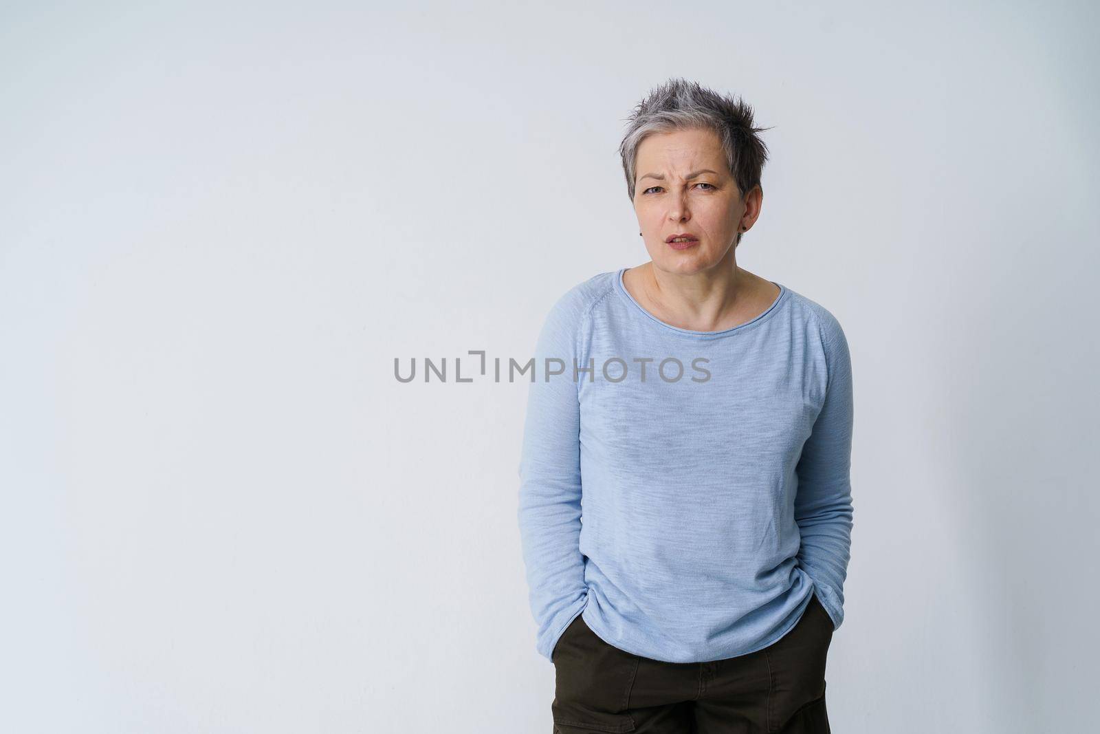 Stressed, angry mature grey haired woman 50s posing frustrated looking at camera with hands in pockets, copy space on left isolated on white background. Mature people healthcare.