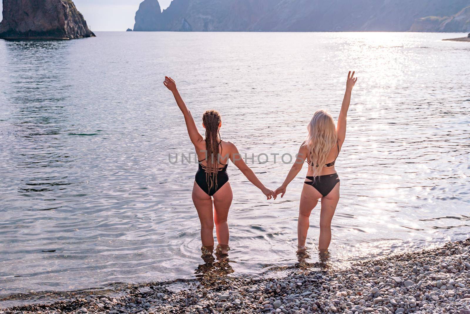 Two beautiful young female friends in black bikinis opened their arms in delight in front of the blue sea and sky by Matiunina