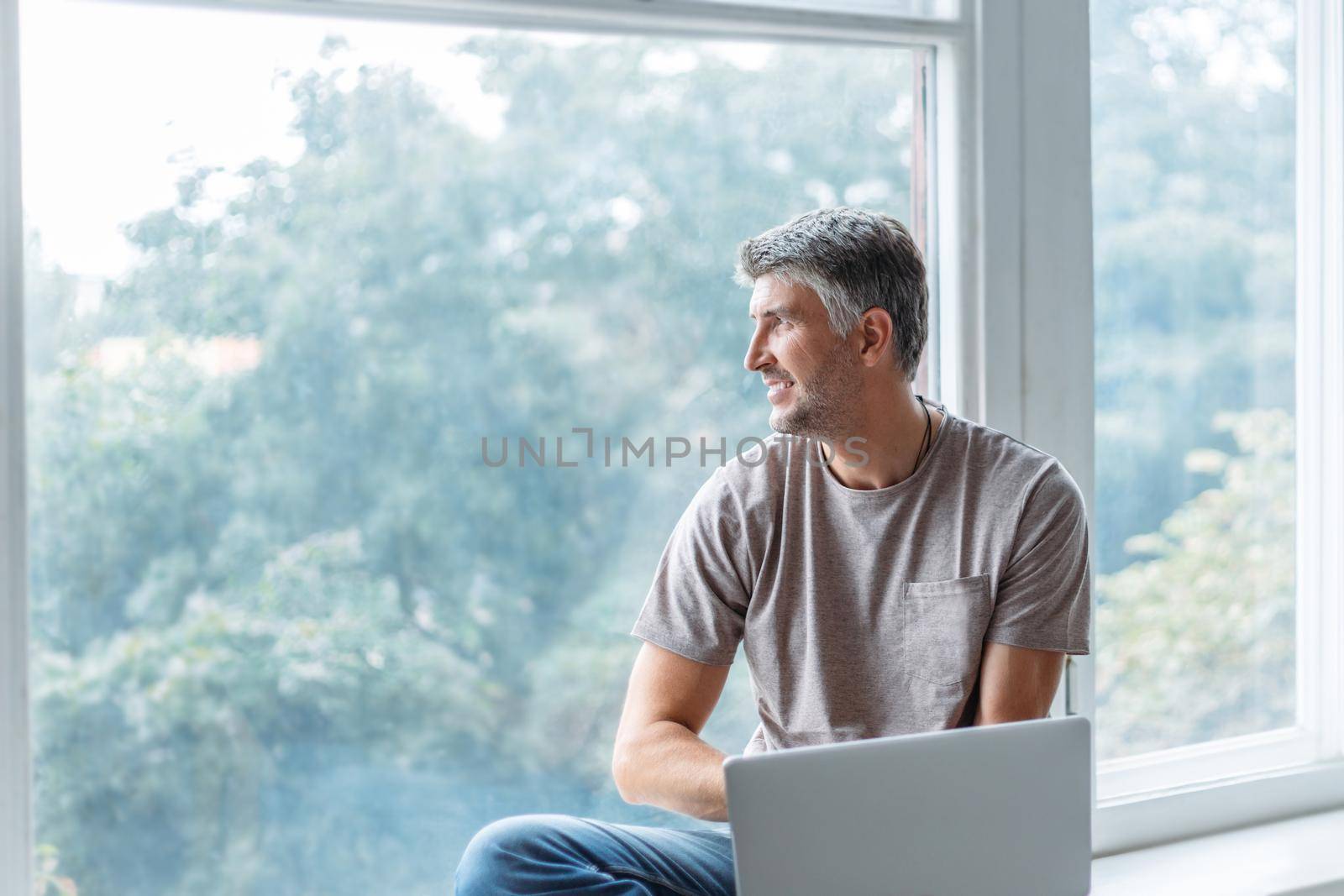man is working on a laptop sitting on the windowsill in the living room. photo with a copy-space.