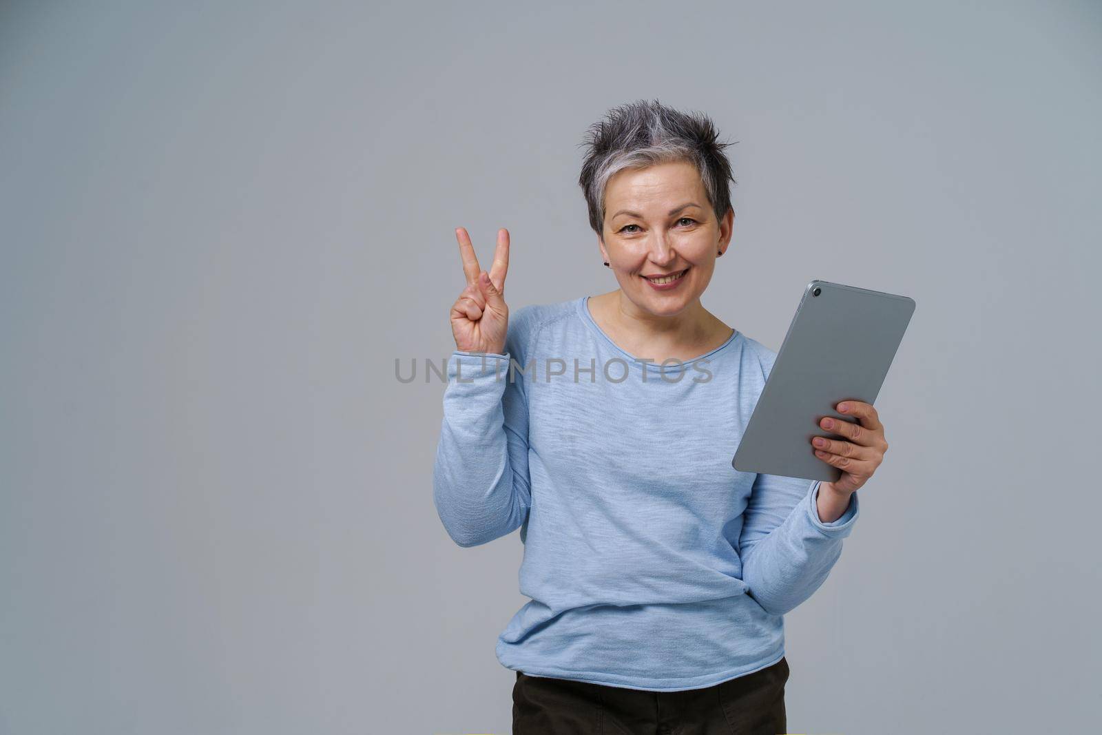 Gesturing V or victory mature grey haired woman in 50s holding digital tablet working or shopping online, checking on social media. Pretty woman in blue blouse isolated on white background.