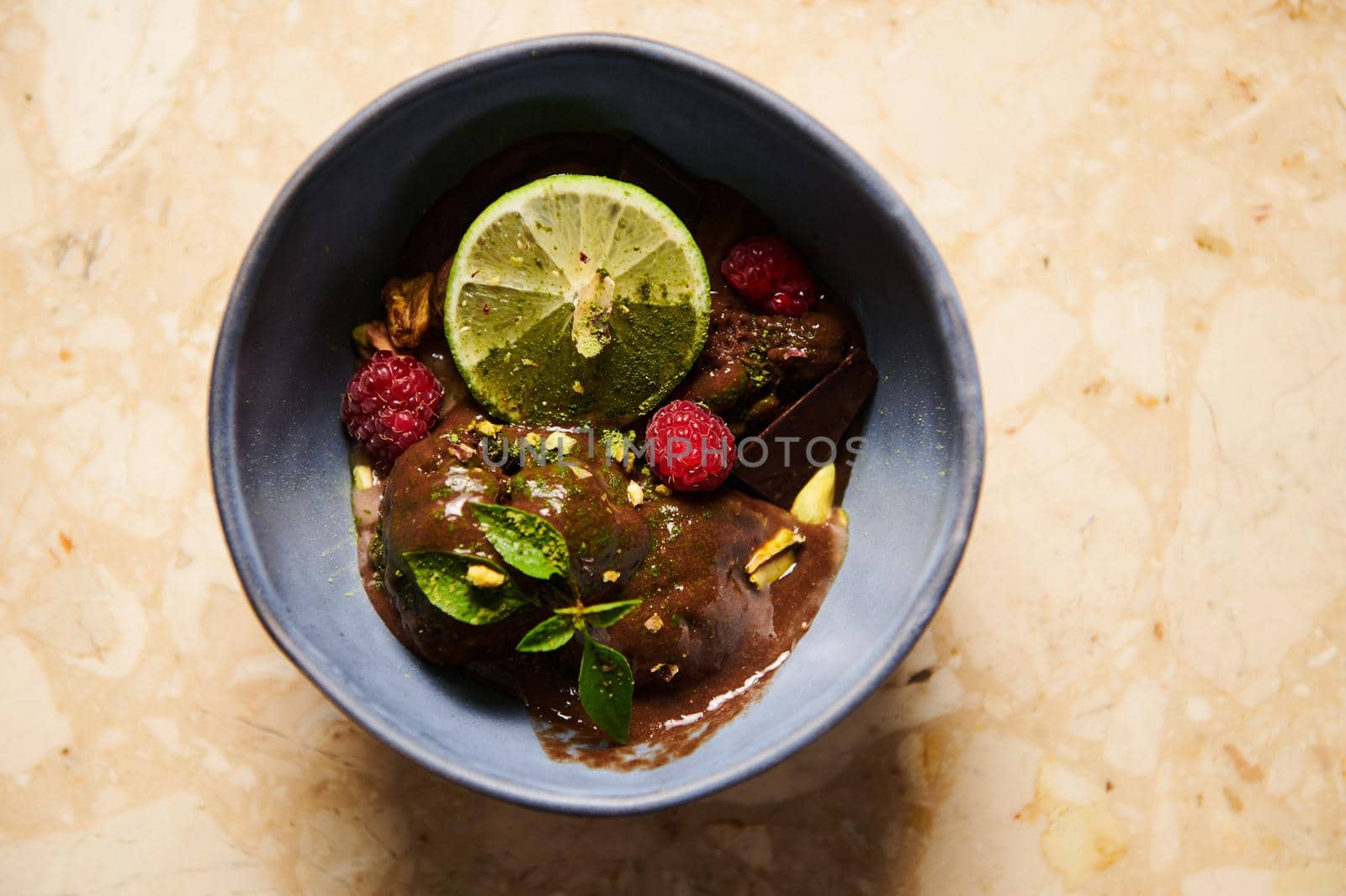 Selective focus on a slice of juicy lime, garnishing a chocolate ice cream, topped with organic cocoa, raspberries and lemon basil leaves, melting in navy blue bowl. Flat lay Still life