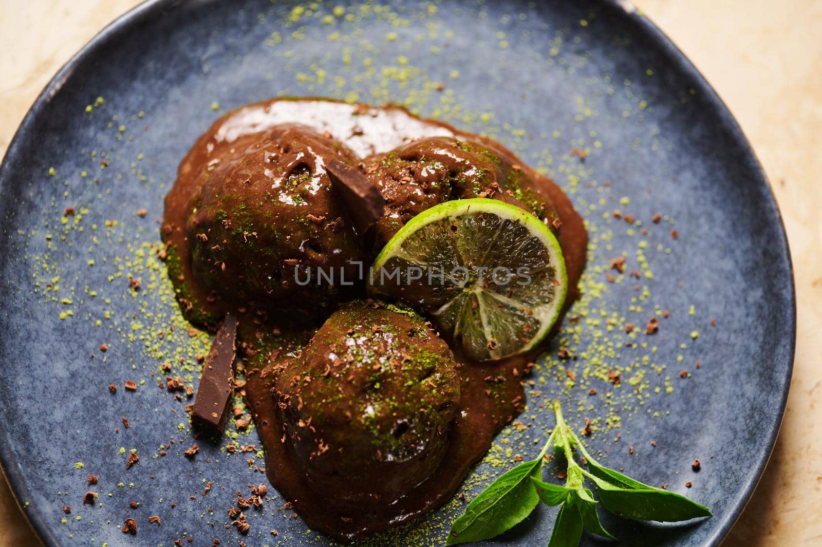 Flat lay. Food still life. Raw vegan chocolate ice cream sorbet balls, garnished with sprinkled green matcha powder, slice of juicy lime and lemon basil leaves on dark blue plate, on marble background