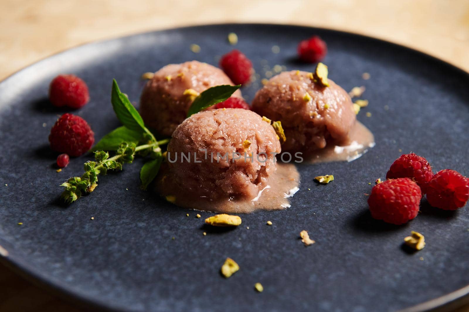 Cropped view of frozen balls with homemade, dairy free berry sorbet, garnished with grated pistachios, raspberries and basil lemon leaves on blue navy plate. Food still life. Selective focus. Close-up