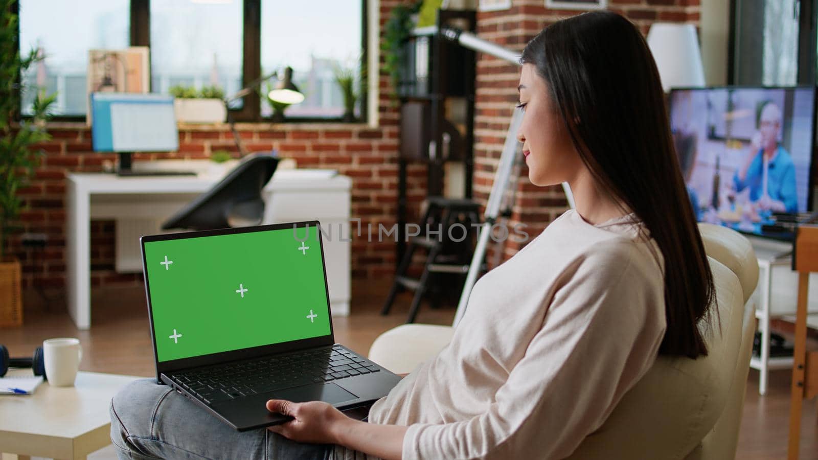 Young adult person sitting on couch while using laptop having chroma key isolated background. Woman sitting on couch inside apartment with portable computer having green screen display.