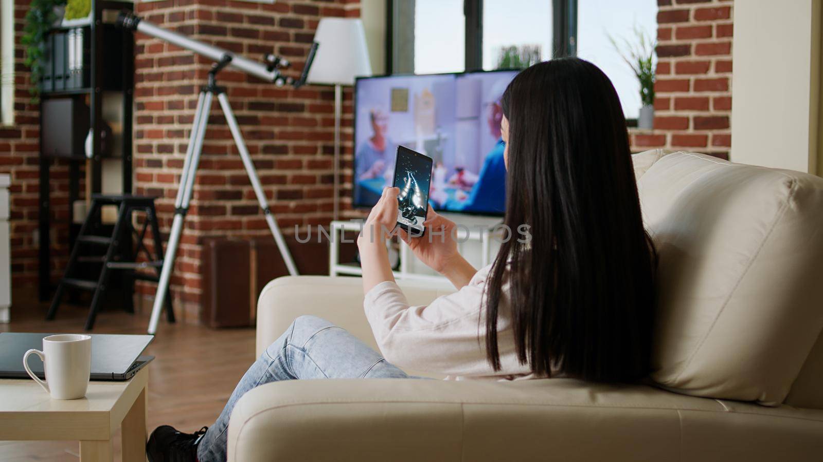 Childish young adult person enjoying mobile gaming while sitting on sofa inside apartment by DCStudio