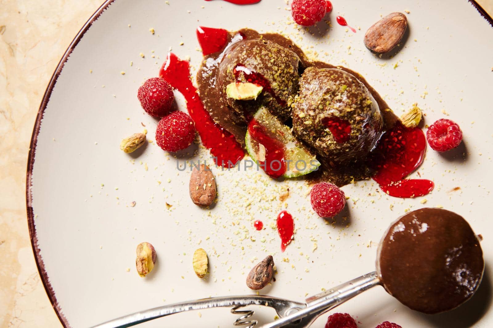 Top view of a metal scoop for sorbet, with homemade, dairy free chocolate ice cream, garnished with grated pistachios, raspberries, cocoa beans and berry jam on a white plate. Food still life