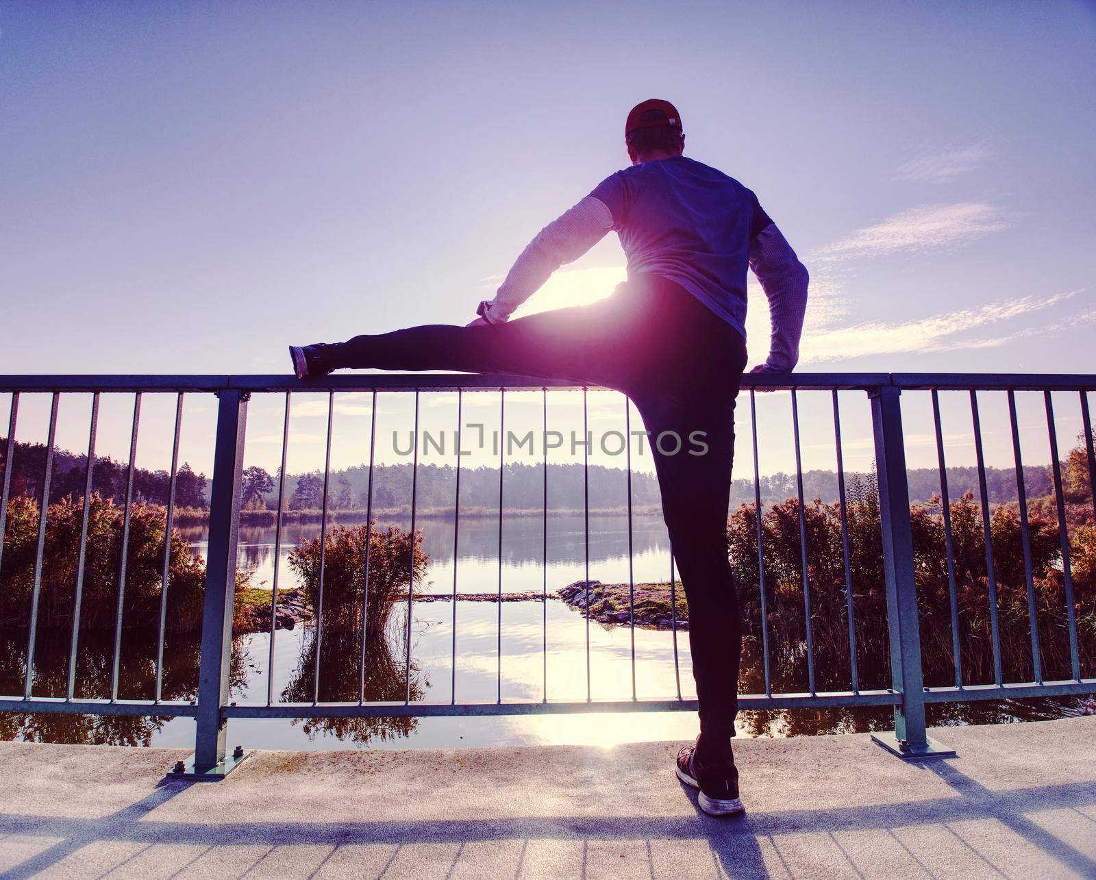 Man stretching legs before going  run outside within sunny morning. Warming up muscles. Outdoor exercising on lake bridge.